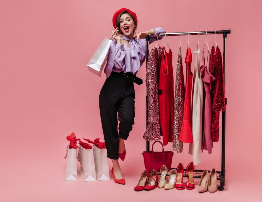 woman-in-black-trousers-and-purple-blouse-laughs-leaning-on-stand-with-elegant-clothes-on-pink-background-min